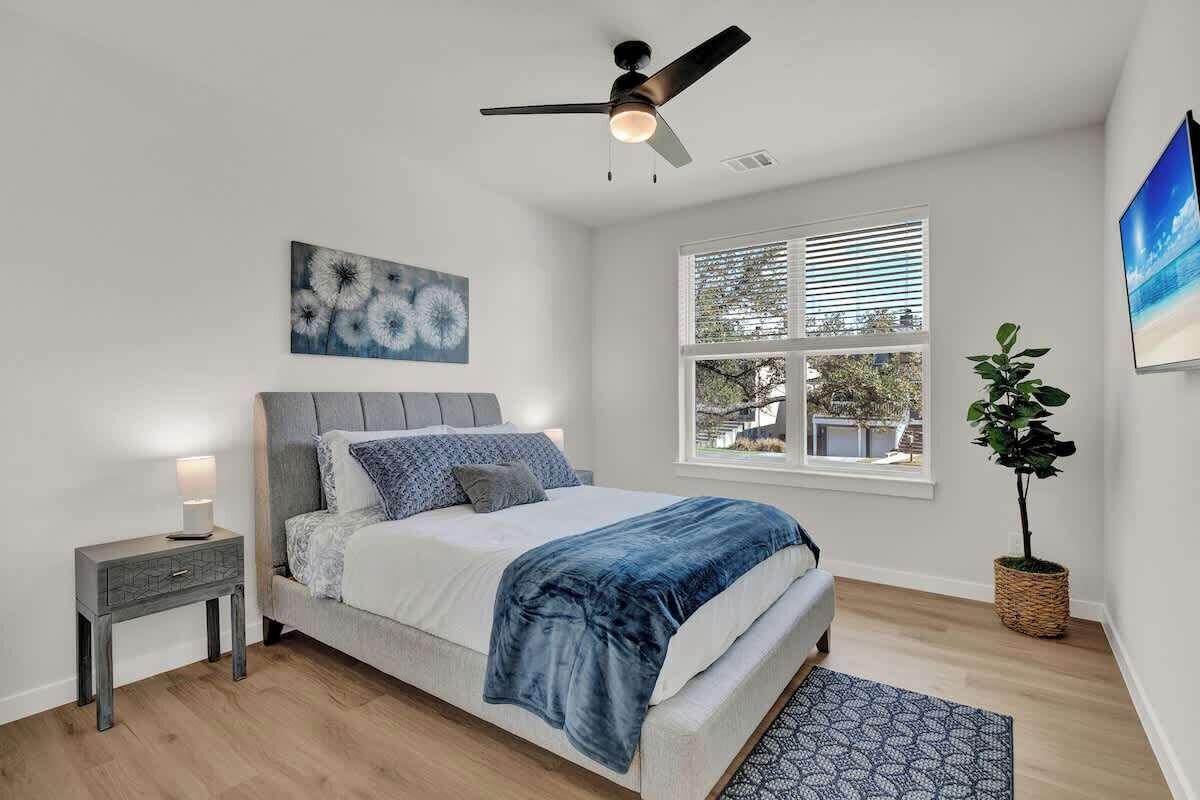 Une chambre à coucher comprenant un lit, une table de chevet, un ventilateur de plafond, une fenêtre avec des stores et une plante en pot.