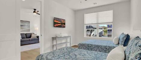 Une chambre avec deux lits, un ventilateur de plafond, une fenêtre avec des stores et un tableau au mur.