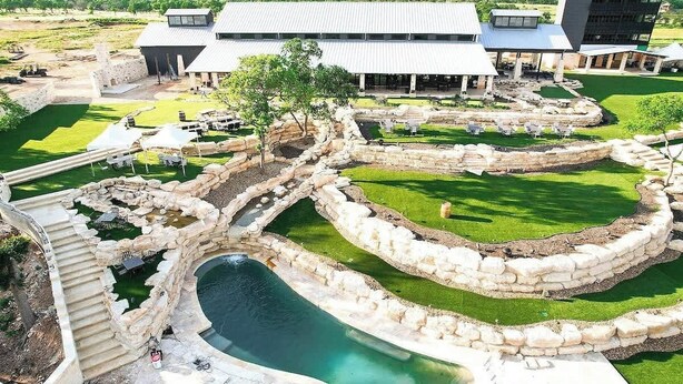 A swimming pool with a stone border, a grassy area, and a building with a flat roof in the background. - Resort at Fredericksburg Unit 1 (Fredericksburg)