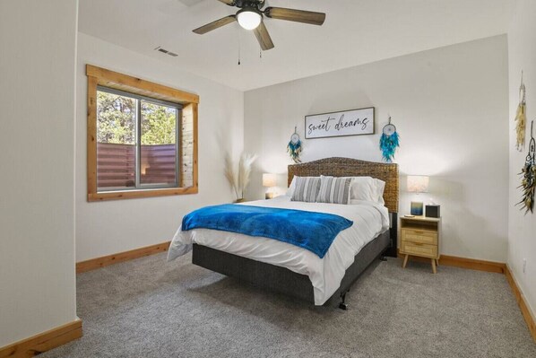 A bedroom with a bed, a ceiling fan, a window, a bedside table with a lamp, and a wall with a dreamcatcher. - Log Cabin | Private Hot Tub | Stunning Lake Views (Divide)