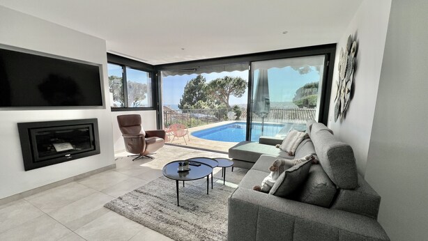 A modern living room with a grey sofa, a TV mounted on the wall, a fireplace, and a pool visible through large windows. - 22 - Villa in Cala de Mijas (La Cala de Mijas)