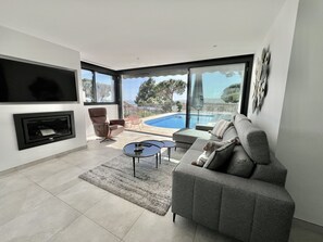 Een moderne woonkamer met een grijze bank, een aan de muur bevestigde televisie, een open haard en een zwembad zichtbaar door grote ramen.
