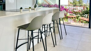 A modern kitchen with a large island, white bar stools, and a view of a balcony with flowers. - 22 - Villa in Cala de Mijas (La Cala de Mijas)