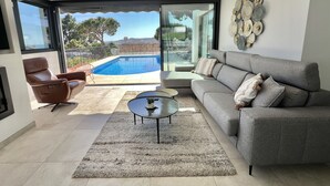 A modern living room with a grey sofa, a brown leather chair, a glass door leading to a pool, and a view of the outdoors. - 22 - Villa in Cala de Mijas (La Cala de Mijas)