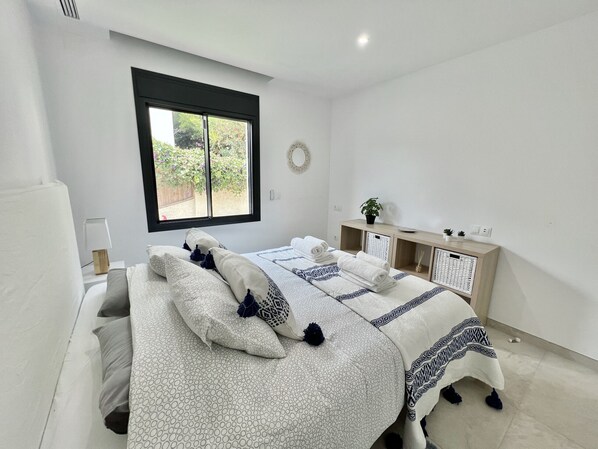 A modern bedroom with a large bed, a bedside table, a small cabinet, and a window with a view of greenery. - 22 - Villa in Cala de Mijas (La Cala de Mijas)
