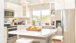 Una cocina moderna con una isla grande, dos taburetes de madera, iluminación colgante, electrodomésticos de acero inoxidable y vistas al exterior.