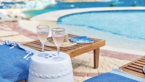Escena junto a la piscina con dos vasos de una bebida de color claro sobre un enfriador blanco, una mesa de madera y vista al océano y edificios al fondo.