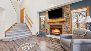 Una sala de estar con chimenea, televisor montado en la pared, escalera y vista al exterior.