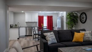 A modern living room with a black sofa, a coffee table, and a dining area with red curtains. - Spacious Basement Apartment (Eagle Mountain)