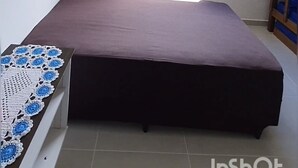 A bedroom with a bed, a window with blinds, a hanging light fixture, and a small wooden shelf.