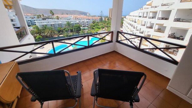 A balcony with two black chairs, a wooden table, and a view of a swimming pool and buildings. - San Marino Studio 7 (Los Cristianos)