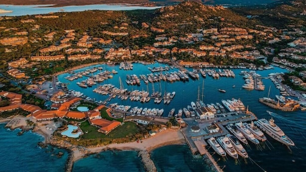 An aerial view of a coastal town with a marina filled with boats, surrounded by residential buildings and natural landscapes. - Porto Rotondo Vista Incantevole sul Mare e Yacht (Olbia)