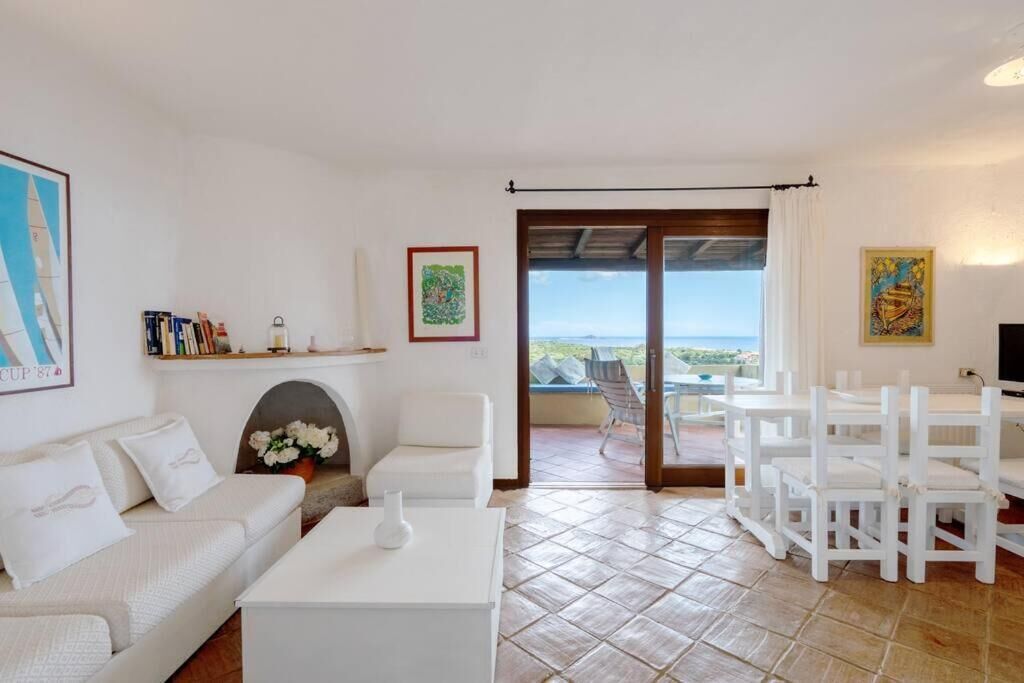 Uma sala de estar ampla com sofá branco, mesa de centro e área de jantar com cadeiras brancas. Há uma lareira com um arranjo de flores, e uma grande janela com vista para o mar.