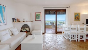 A spacious living room with a white sofa, a coffee table, and a dining area with white chairs. There is a fireplace with a flower arrangement, and a large window offering a view of the sea. - Porto Rotondo Vista Incantevole sul Mare e Yacht (Olbia)
