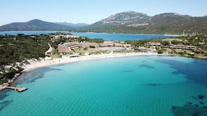 Luftaufnahme eines Küstenresorts mit Strand, klarem türkisblauem Wasser und einem gebirgigen Hintergrund.