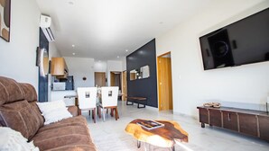 Uma sala de estar moderna com um sofá marrom, uma mesa de centro de madeira, uma TV e uma área de jantar com cadeiras brancas.