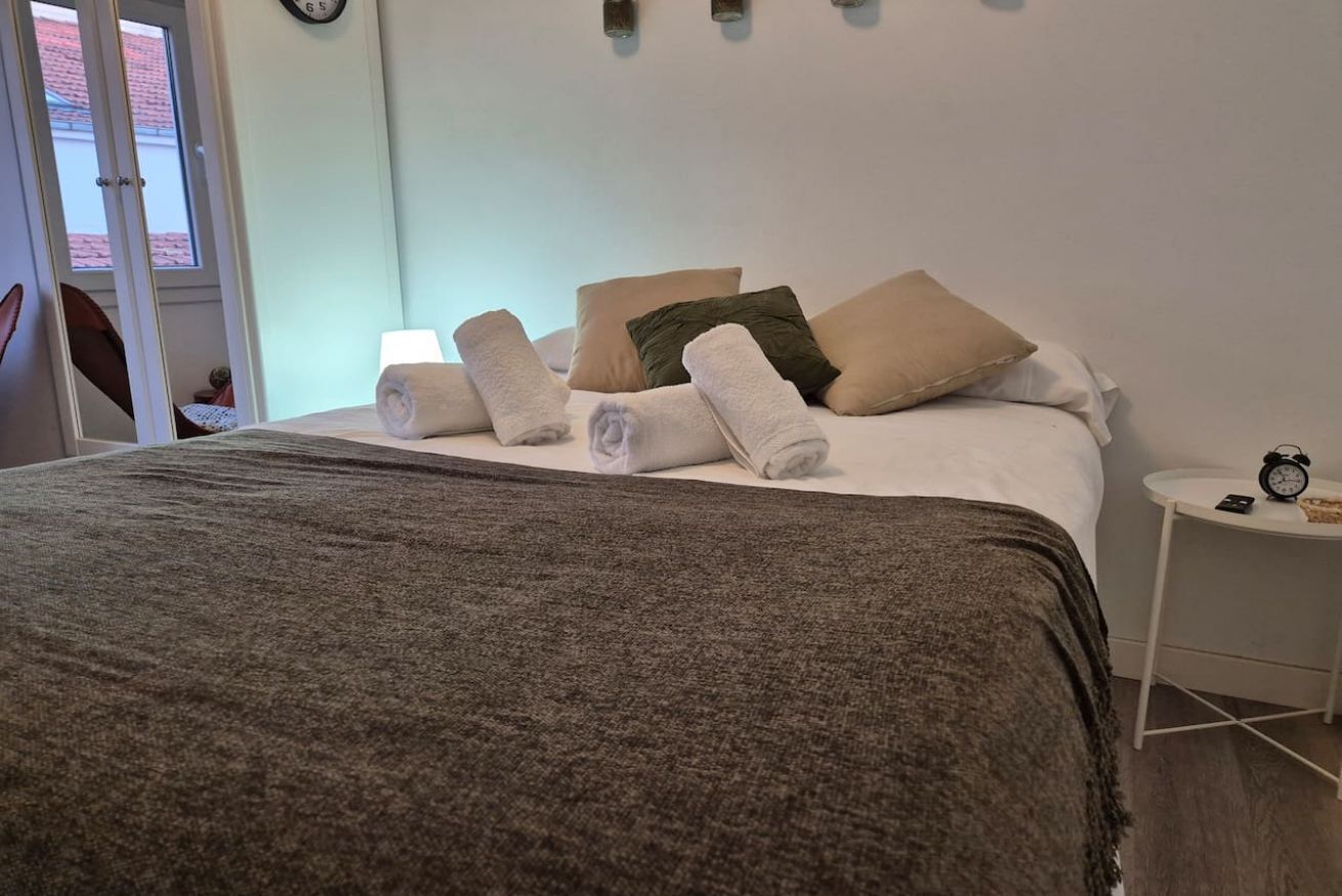 Een net opgemaakt bed met opgerolde handdoeken, een nachtkastje met een klok, en uitzicht op een gebouw met rode dakpannen.