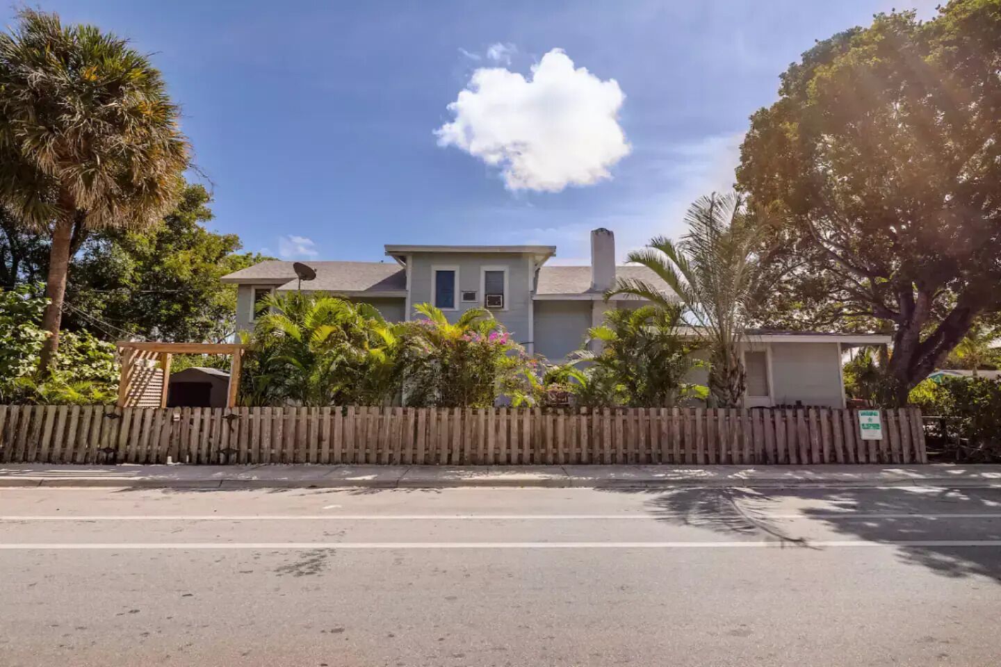 A two-story house with a fenced yard, surrounded by lush greenery and palm trees.