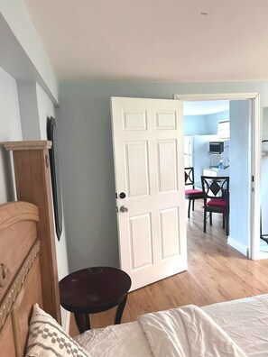Ein Schlafzimmer mit einem Bett, einem hölzernen Kopfteil, einem kleinen runden Tisch und einer weißen Tür, die in einen Essbereich mit Tisch und Stühlen führt.