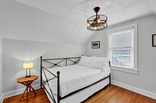 A bedroom with a bed, a bedside table with a lamp, a window with blinds, and a ceiling light fixture.