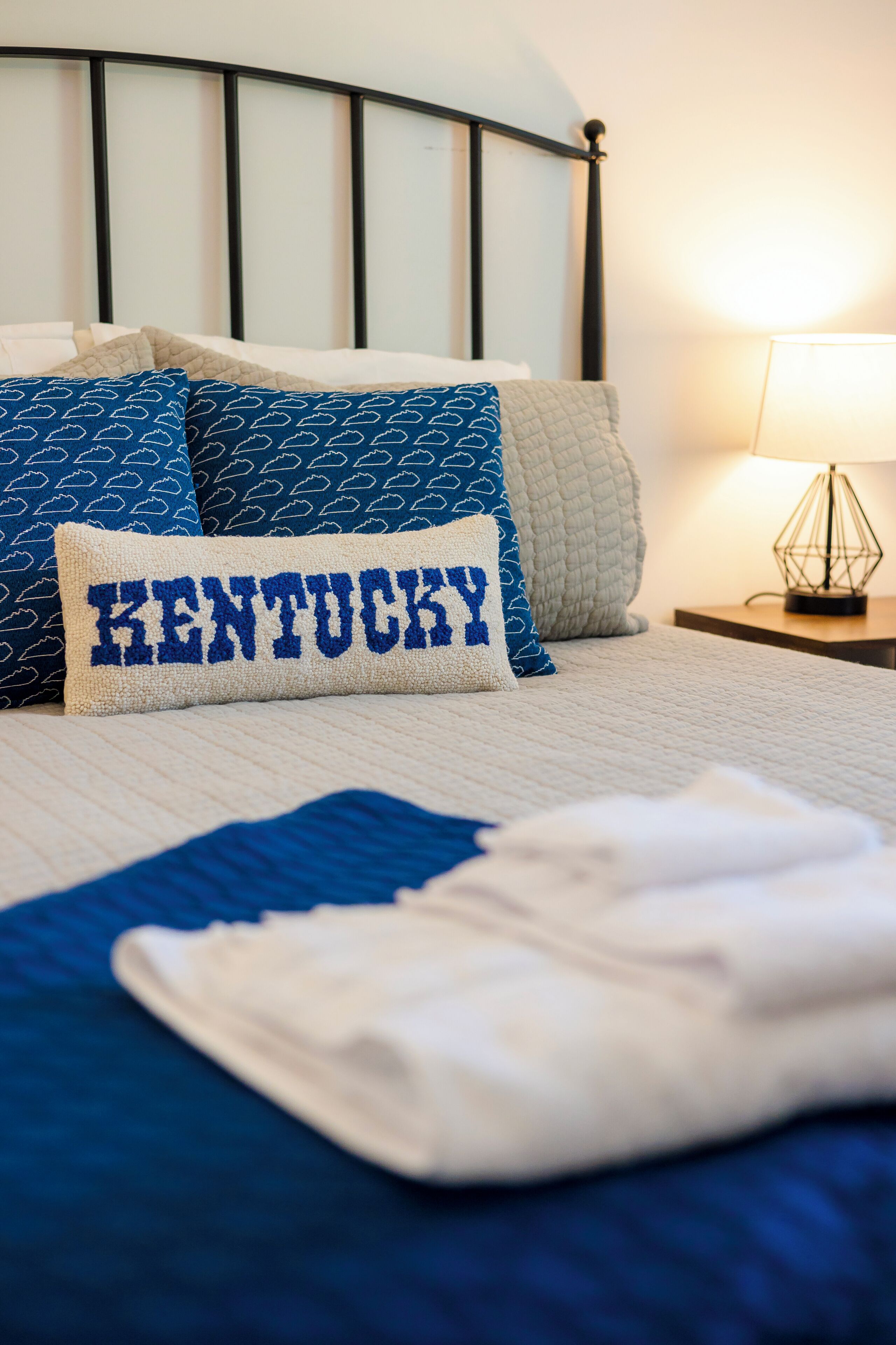 A neatly made bed with a pillow labeled "KENTUCKY," a bedside lamp, and a quilted blanket.