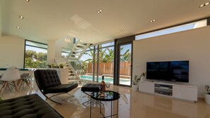 Un salon moderne avec une grande télévision, une table basse en verre, un canapé noir et un escalier menant à un balcon.