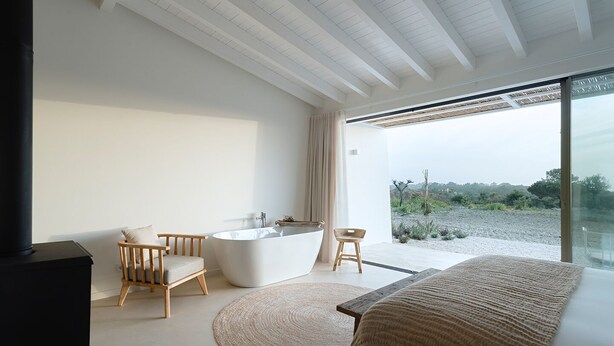 A modern bathroom with a freestanding bathtub, a wooden chair, and a large window offering a view of the outdoors. - Amaria (Aljezur)