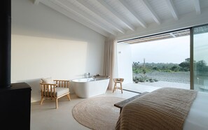 A modern bathroom with a freestanding bathtub, a wooden chair, and a large window offering a view of the outdoors. - Amaria (Aljezur)