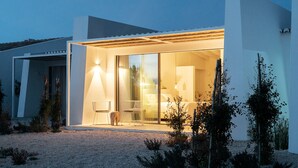 A modern house with a glass door entrance, a visible bedroom with a bed and a nightstand, and a small outdoor seating area with two chairs. - Amaria (Aljezur)