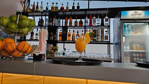 Uma área de bar com diversos frascos de bebidas, uma cesta de frutas com maçãs verdes e laranjas, e um copo de suco de laranja.