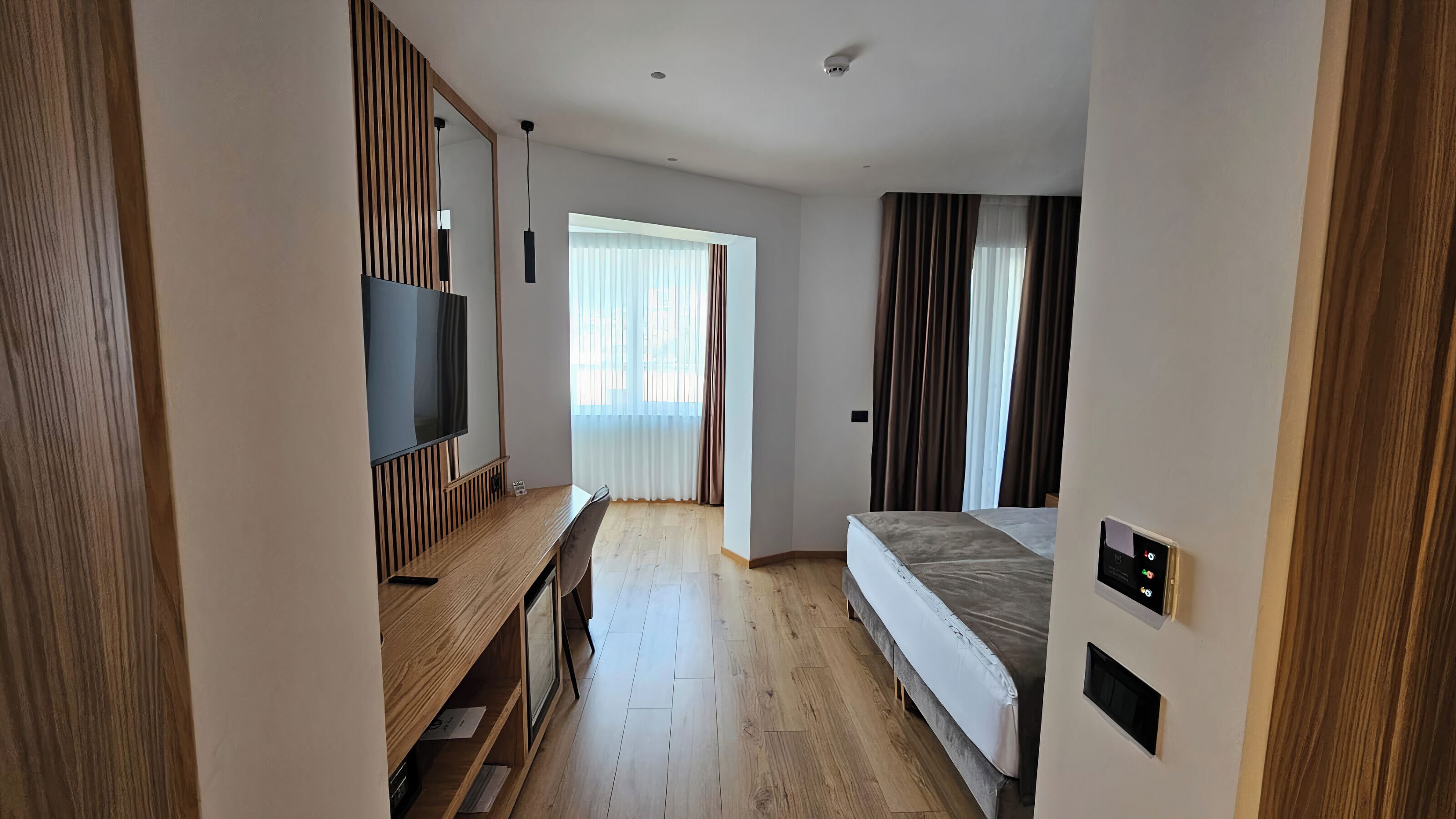 Una habitación de hotel moderna con un escritorio de madera, un televisor de pantalla plana, una cama y un gran ventanal con cortinas.