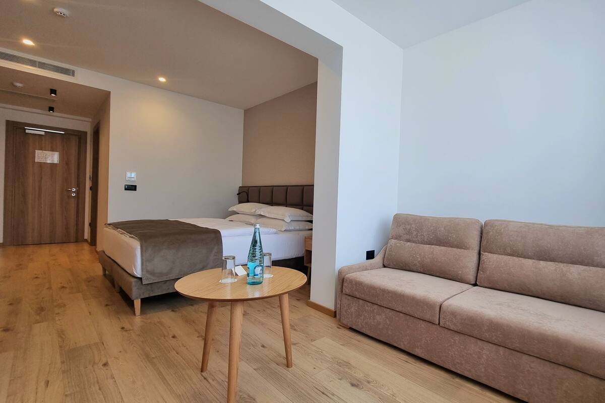 A hotel room with a bed, a sofa, a small round table with a bottle and glasses, and a door with a sign.