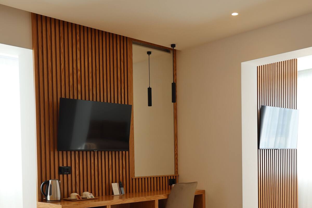 A modern hotel room with a wooden floor, a bed, a TV mounted on a wooden panel wall, a desk with a chair, and a small refrigerator.