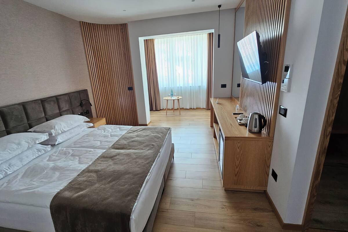 A hotel room with a large bed, a wooden desk with a flat-screen TV, and a small table with a kettle and cups.