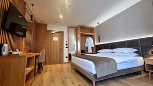 Quarto de hotel moderno com uma secretária de madeira, uma cama com várias almofadas, um roupão pendurado e uma televisão na parede.