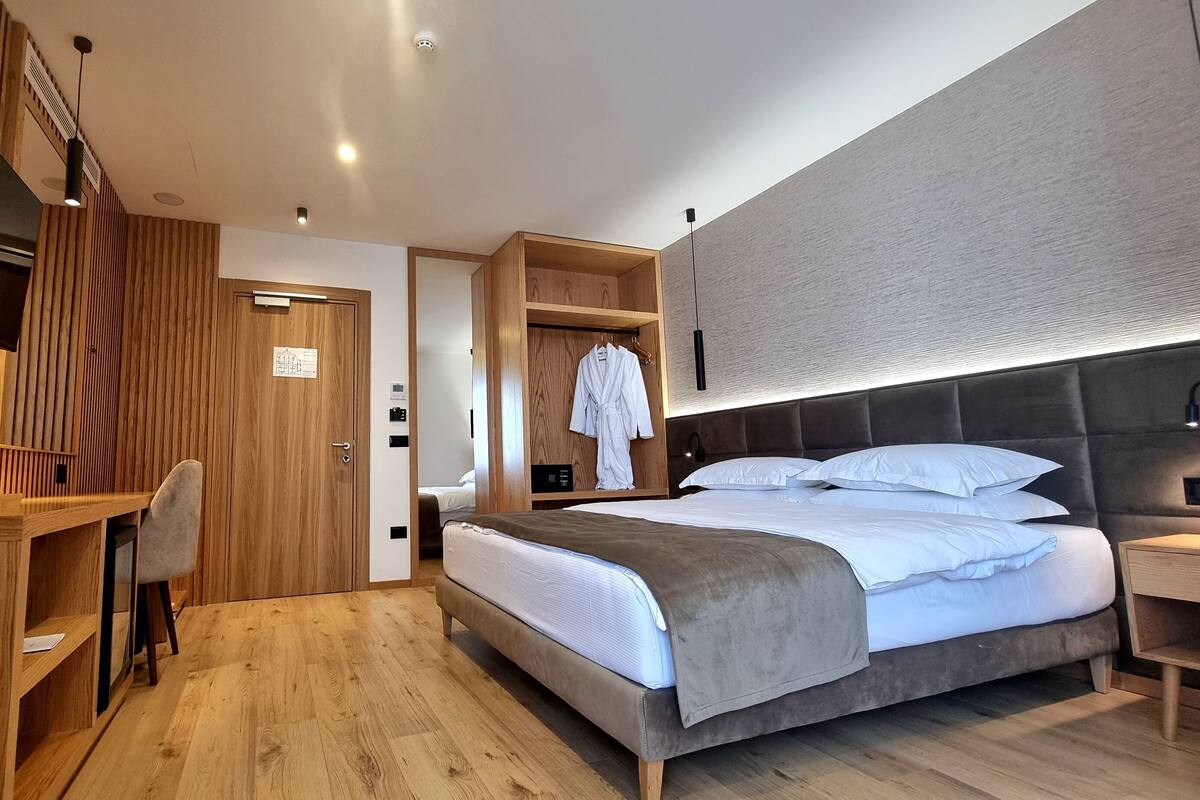 A modern hotel room with a wooden desk, a bed with multiple pillows, a robe hanging, and a TV mounted on the wall.
