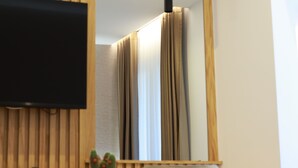 Quarto de hotel moderno com parede de painel de madeira, uma TV de tela plana acima de uma escrivaninha, uma planta em vaso e um espelho grande refletindo o quarto.
