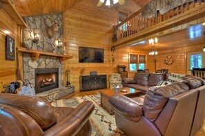 A log cabin interior with a stone fireplace, leather sofas, and a balcony.