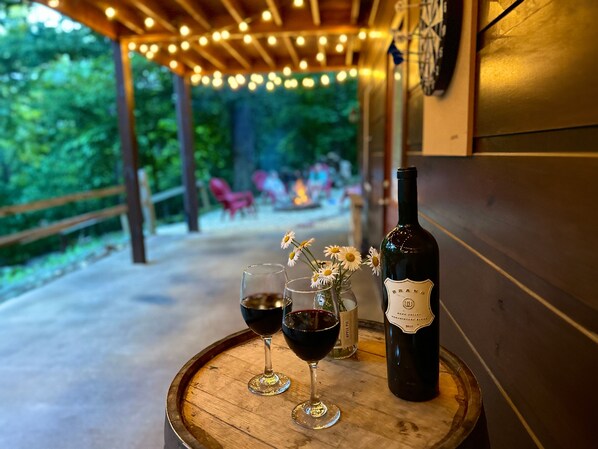 A wooden barrel table with two wine glasses and a bottle of wine, set outdoors under a wooden pergola with string lights.