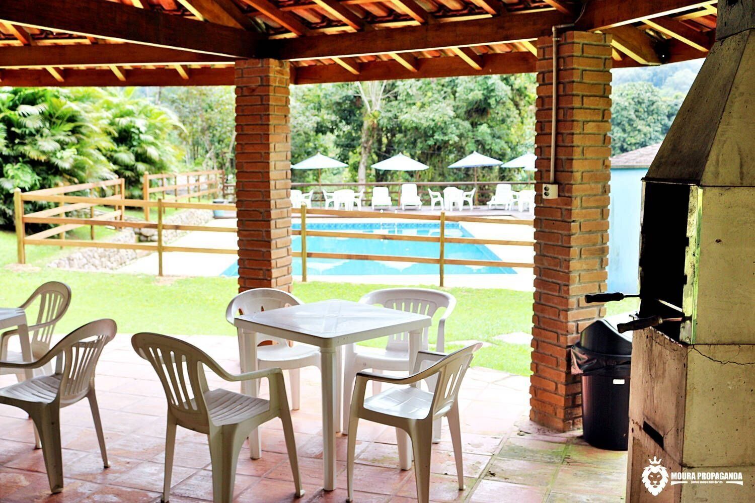 Um pátio externo com área de churrasqueira de tijolos, piscina e cadeiras e mesas de plástico branco.