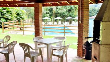 Um pátio externo com área de churrasqueira de tijolos, piscina e cadeiras e mesas de plástico branco.