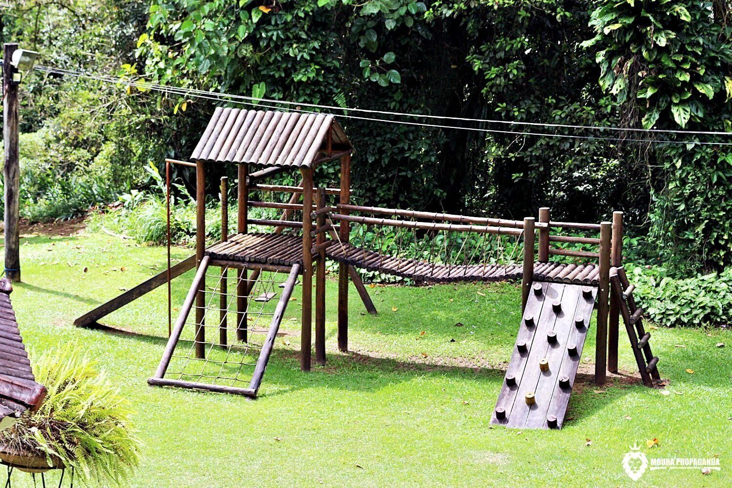 A wooden play structure with a roof and a slide in a grassy area with dense foliage in the background.