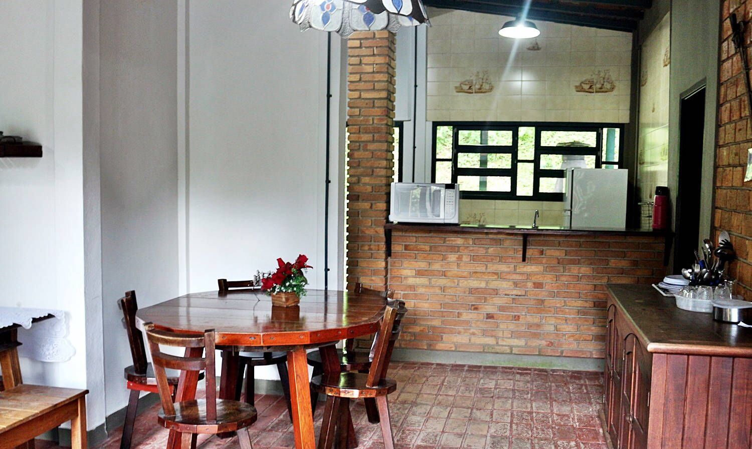 A dining area with a wooden table and chairs, a flower vase, and a brick wall separating it from the kitchen area with a microwave and sink.