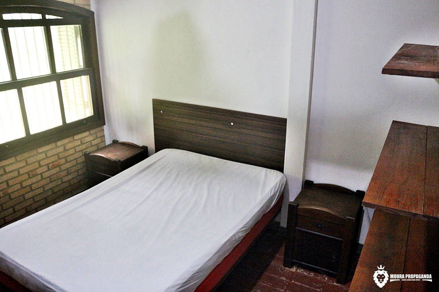 Una cama individual con cabecera de madera, una mesita de noche pequeña y una ventana con persianas.