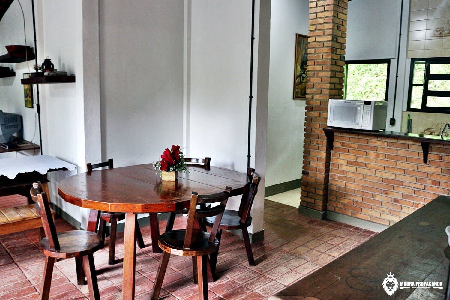 Un comedor con una mesa y sillas de madera, un jarrón con flores rojas y una pared de ladrillo con una ventana.