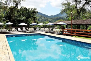 Piscina con sillas de descanso y sombrillas, rodeada de vegetación y montañas.