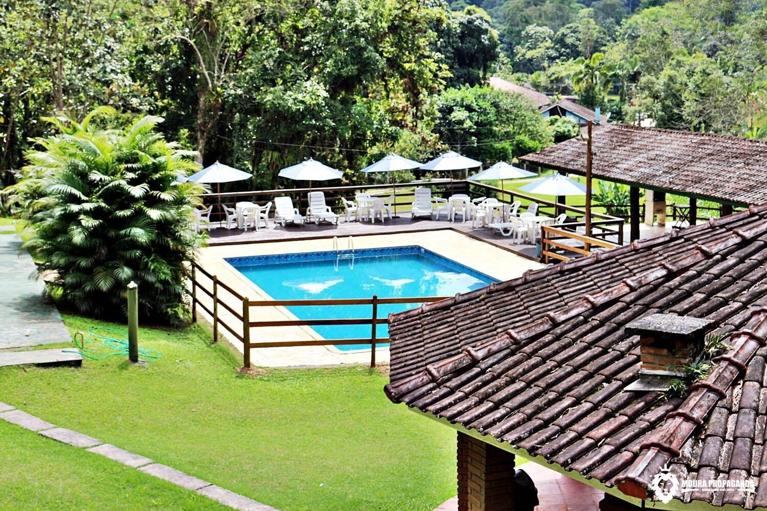 A swimming pool with a wooden deck, surrounded by white lounge chairs and umbrellas, set against a lush green landscape.