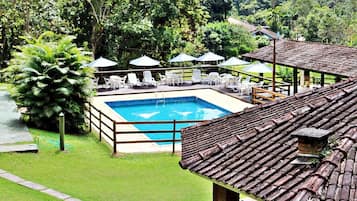 A swimming pool with a wooden deck, surrounded by white lounge chairs and umbrellas, set against a lush green landscape.