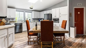 A modern kitchen with white cabinets, a dining table with chairs, a refrigerator, and a dishwasher. - Cowboys & Roses - Wine Country Home w/Game Room, Hot Tub & Views - Dogs Welcome! (Cornville)