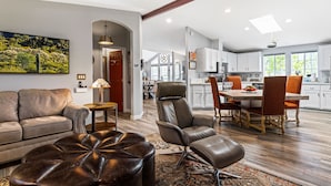 A spacious living room with a leather sofa, ottoman, and leather chairs, a dining area with a wooden table and chairs, and a kitchen with white cabinets and a window. - Cowboys & Roses - Wine Country Home w/Game Room, Hot Tub & Views - Dogs Welcome! (Cornville)